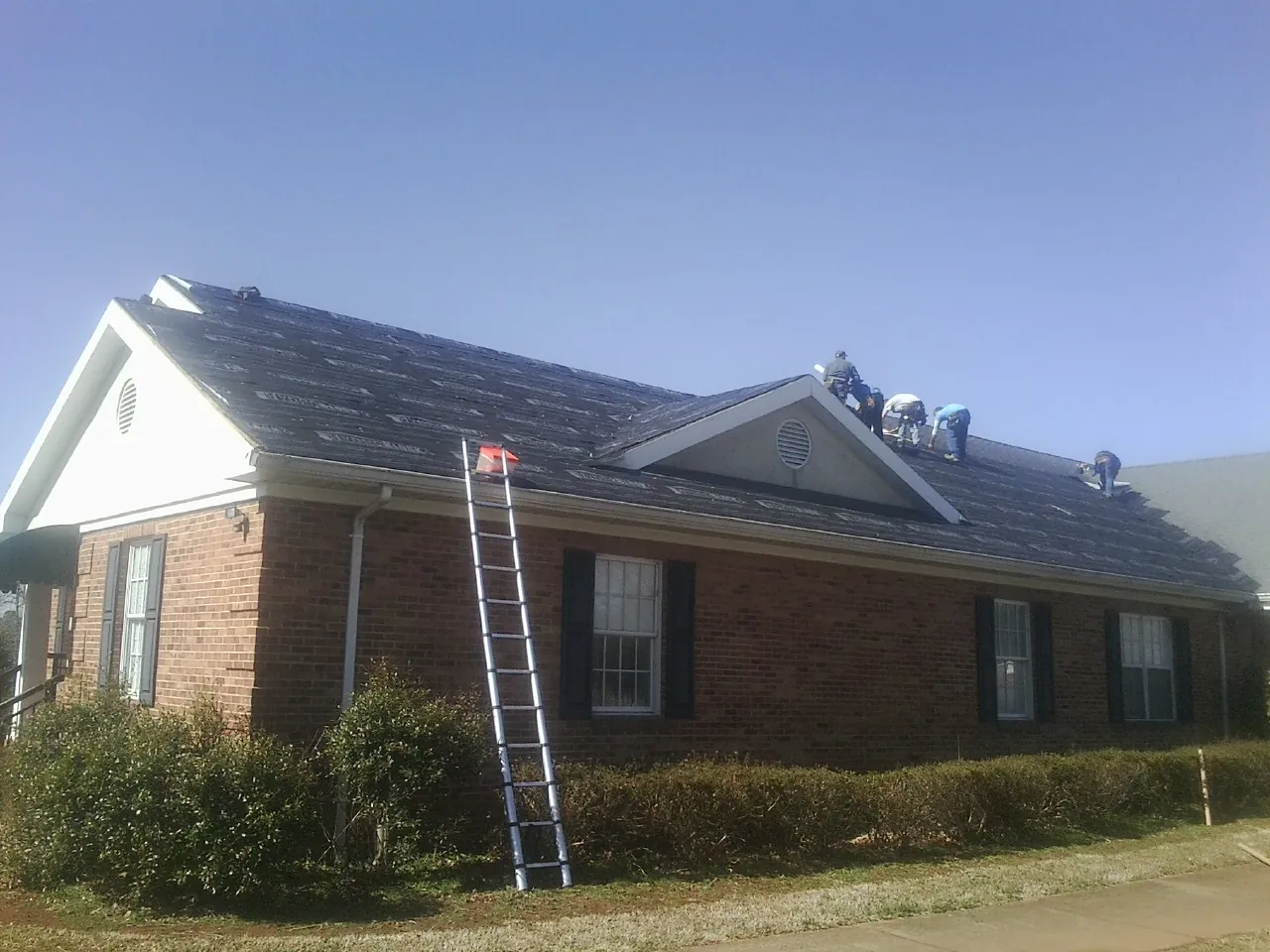 Crew tearing shingles off a brick ranch roof with a ladder set — Emergency Roof Repair work by our crew in Hartfield