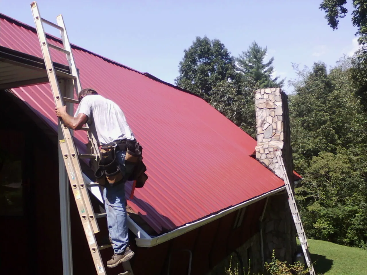 Roofer on a ladder beside a red metal roof and stone chimney — Soffit & Fascia Repair by Roofing Brothers in Hartfield